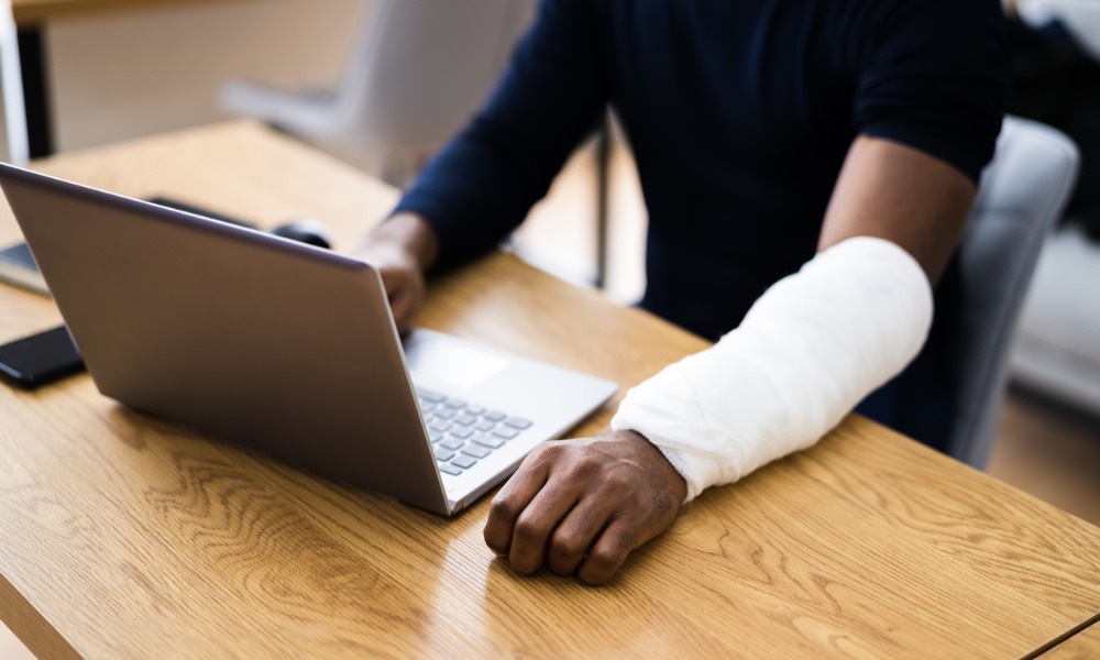 A remote employee types on a laptop with a bandaged arm after filing a claim for workers’ compensation while working from home.